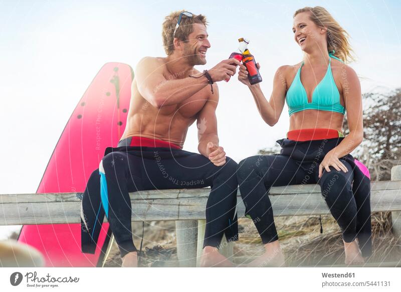 Happy couple with surfboard having a drink on the beach laughing Laughter drinking beaches twosomes partnership couples positive Emotion Feeling Feelings