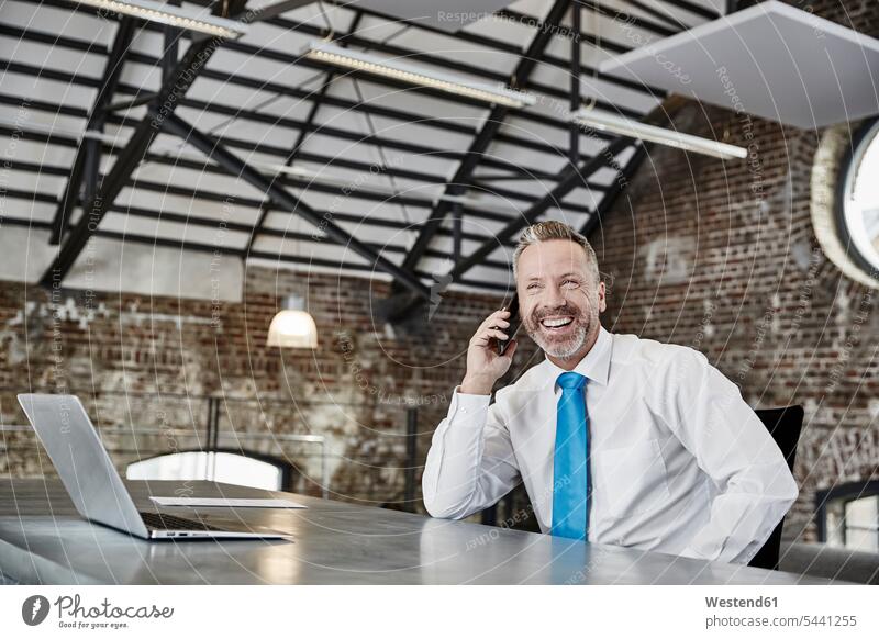 Happy businessman on cell phone sitting at table in a loft on the phone call telephoning On The Telephone calling Seated mobile phone mobiles mobile phones