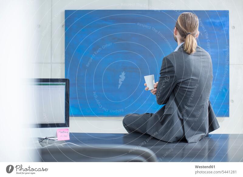 Businessman in office sitting on desk looking at a blue painting Seated Painted Image Painted Images Fine Art Painting paintings picture pictures Business man