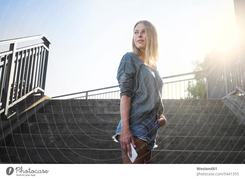 Blond young woman with smartphone standing at backlight in front of stairs Smartphone iPhone Smartphones females women blond blond hair blonde hair Backlit