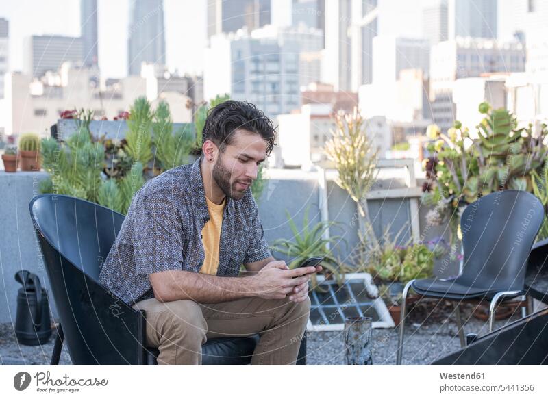 Young man sitting on rooftop terrace checking smart phone messages men males chair chairs mobile phone mobiles mobile phones Cellphone cell phone cell phones