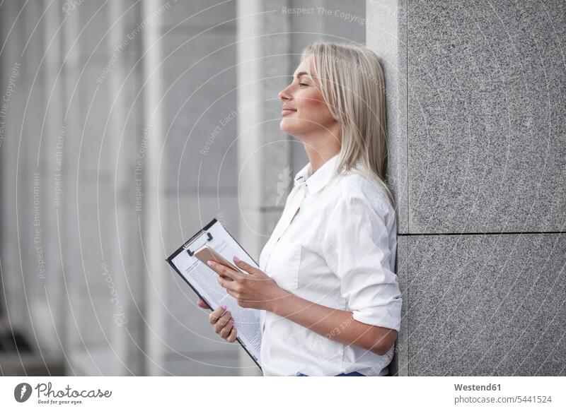 Smiling businesswoman with clipboard and cell phone leaning against a wall businesswomen business woman business women mobile phone mobiles mobile phones