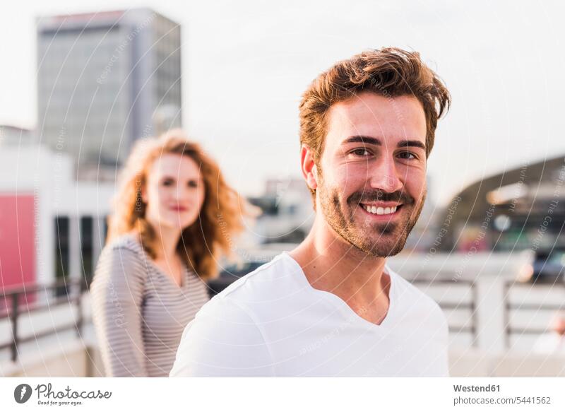 Portrait of happy young man with girlfriend in the background at sunset men males happiness sunsets sundown portrait portraits Adults grown-ups grownups adult