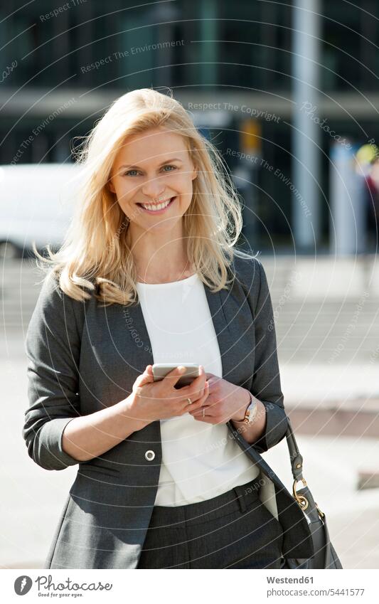 Portrait of smiling blond businesswoman with cell phone and handbag businesswomen business woman business women portrait portraits business people