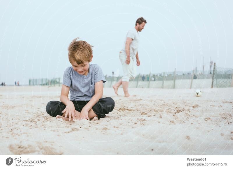 Smiling little boy sitting on the sand on the beach while his father playing with ball in the background beaches pa fathers daddy dads papa son sons manchild