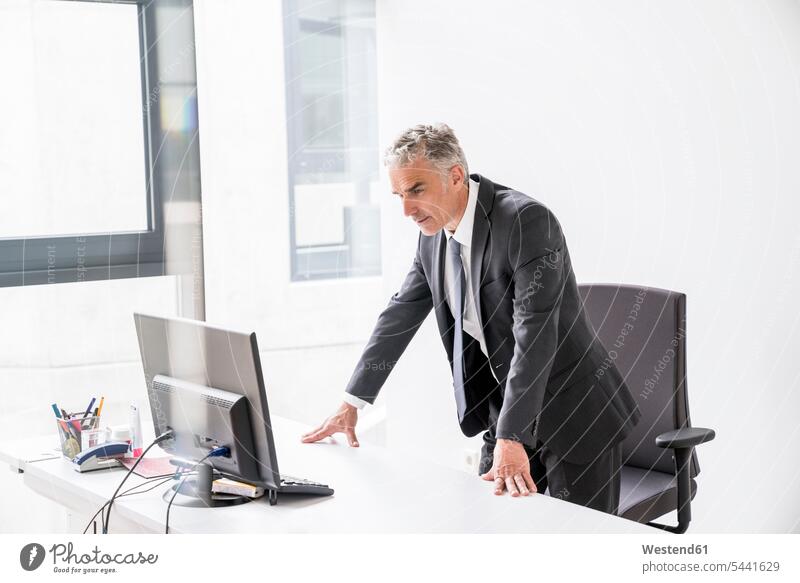 Mature businessman standing in office, looking at computer Businessman Business man Businessmen Business men manager managers offices office room office rooms