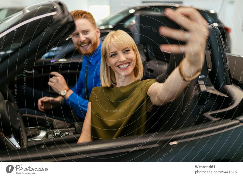 Couple taking selfies, sitting in convertible in car dealership automobile Auto cars motorcars Automobiles Selfie Selfies couple twosomes partnership couples