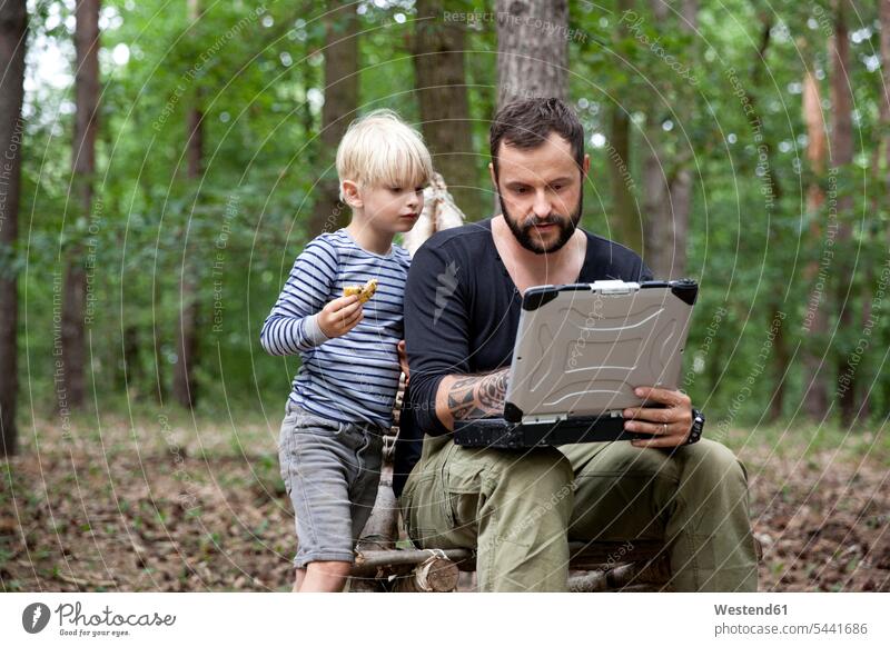 Father and son sitting on self-made wooden chair in forest using laptop Laptop Computers laptops notebook man men males sons manchild manchildren woods forests