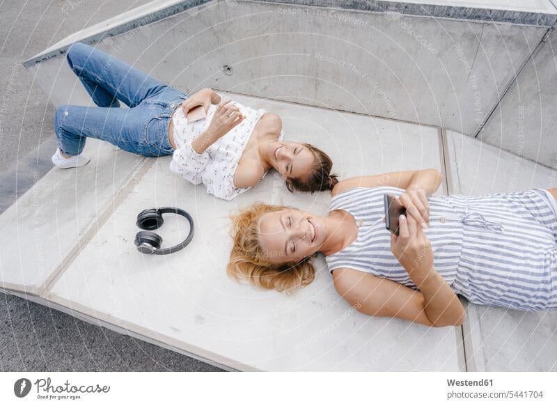 Two happy young women with cell phone and headphones in a skatepark female friends Skateboard Park skate park happiness woman females headset mobile phone
