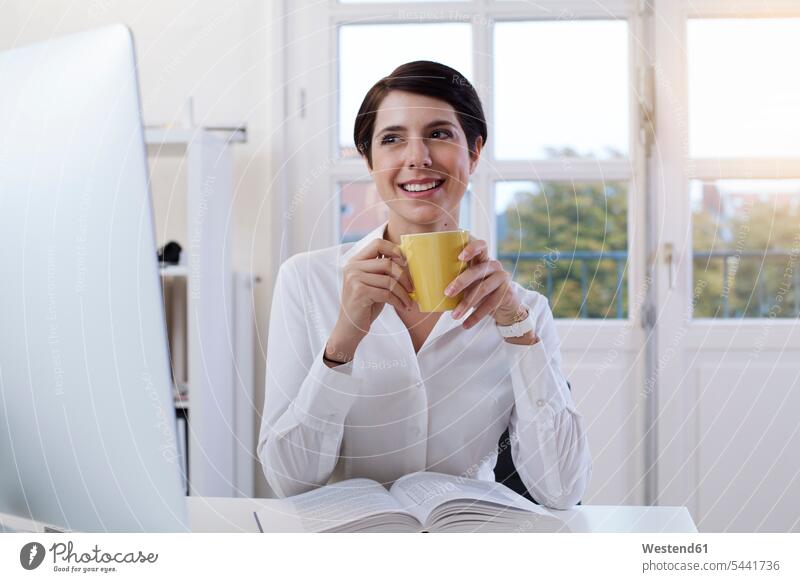 Smiling woman holding cup of coffee at desk in office offices office room office rooms females women Coffee smiling smile workplace work place place of work
