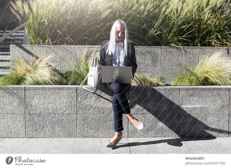Young businesswoman sitting on a wall in the city using laptop businesswomen business woman business women Laptop Computers laptops notebook females Seated