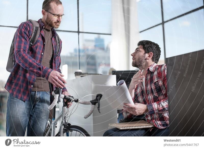 Casual man with bike in modern office talking to colleague men males bicycle bikes bicycles colleagues offices office room office rooms Adults grown-ups
