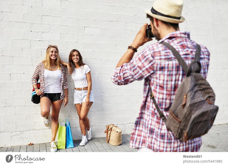 Man taking picture of two young women standing at white wall with bags smiling smile friends mate photographing friendship walls Spain female tourist happiness