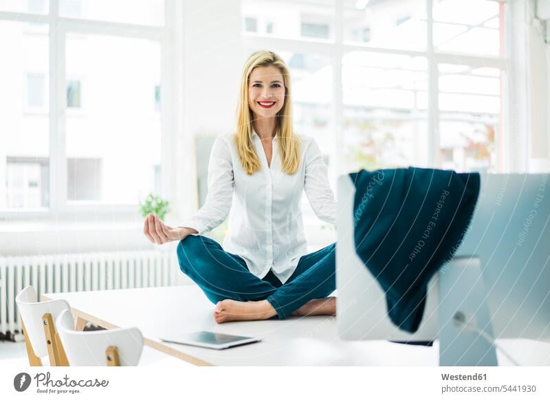 Smiling businesswoman sitting on desk in office practicing yoga businesswomen business woman business women Office Offices smiling smile business people