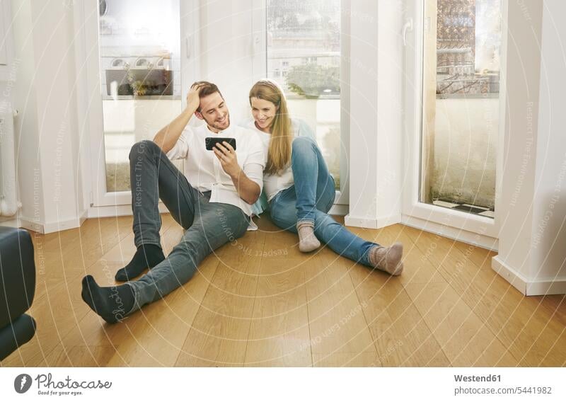 Smiling couple sitting on floor looking at cell phone mobile phone mobiles mobile phones Cellphone cell phones smiling smile twosomes partnership couples