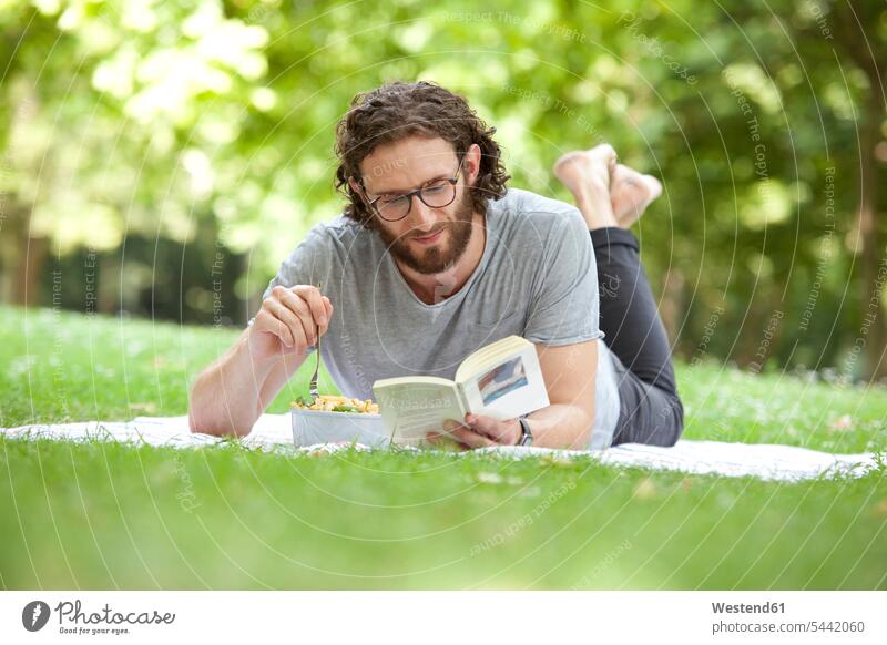 Man reading book on blanket in a park while eating noodle salad man men males books Adults grown-ups grownups adult people persons human being humans