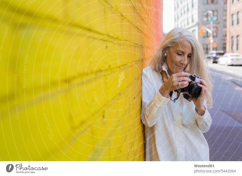 Mature woman with camera leaning against wall females women Adults grown-ups grownups adult people persons human being humans human beings cameras standing