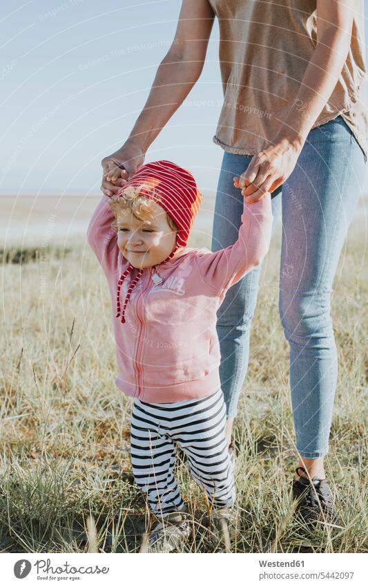 Smiling toddler holding mother's hands on a meadow mommy mothers ma mummy mama daughter daughters parents family families people persons human being humans