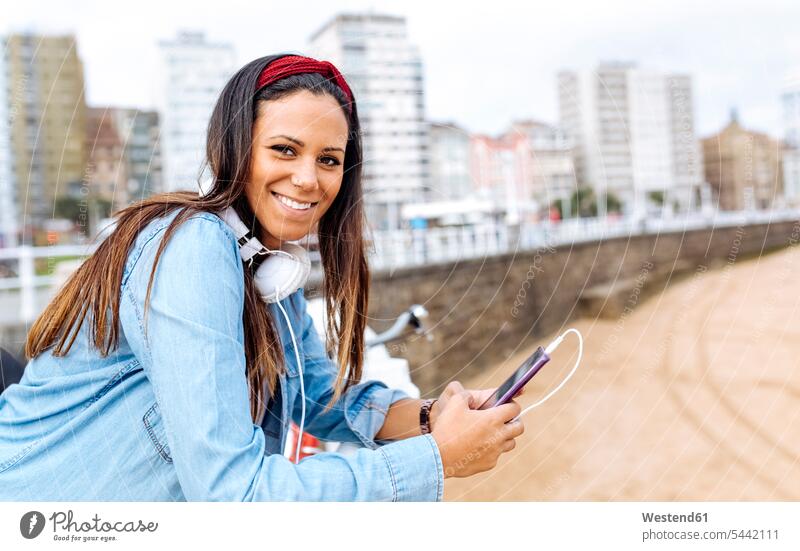 Spain, Gijon, smiling young woman with cell phone and headphones at waterfront promenade smile females women headset mobile phone mobiles mobile phones