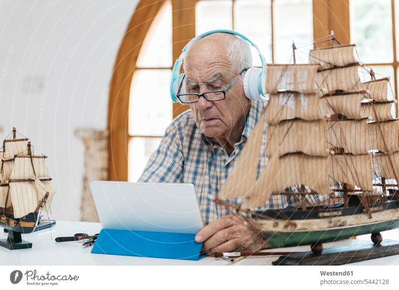 Senior man wearing headphones working on model ship on table with tablet headset senior men senior man elder man elder men senior citizen males model boat