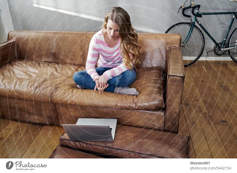 Blond woman sitting on leather couch using tablet females women laptop Laptop Computers laptops notebook settee sofa sofas couches settees Adults grown-ups