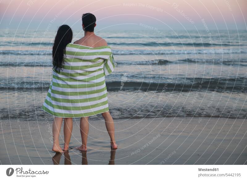 Back view of young couple on the beach wrapped together in towel watching sunset twosomes partnership couples sunsets sundown beaches people persons human being