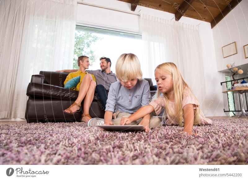 Brother and sister using tablet on carpet in living room with parents in background brother brothers sisters use carpets rug rugs digitizer Tablet Computer