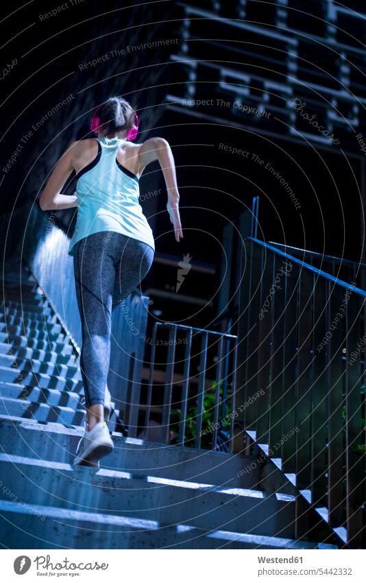 Young woman with pink headphones running upstairs in modern urban setting at night exercising exercise training practising jogger joggers female jogger fit