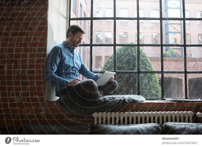 Mature man sitting on window sill, using laptop Laptop Computers laptops notebook working At Work Seated home at home windowsill sills window sills Window Cill