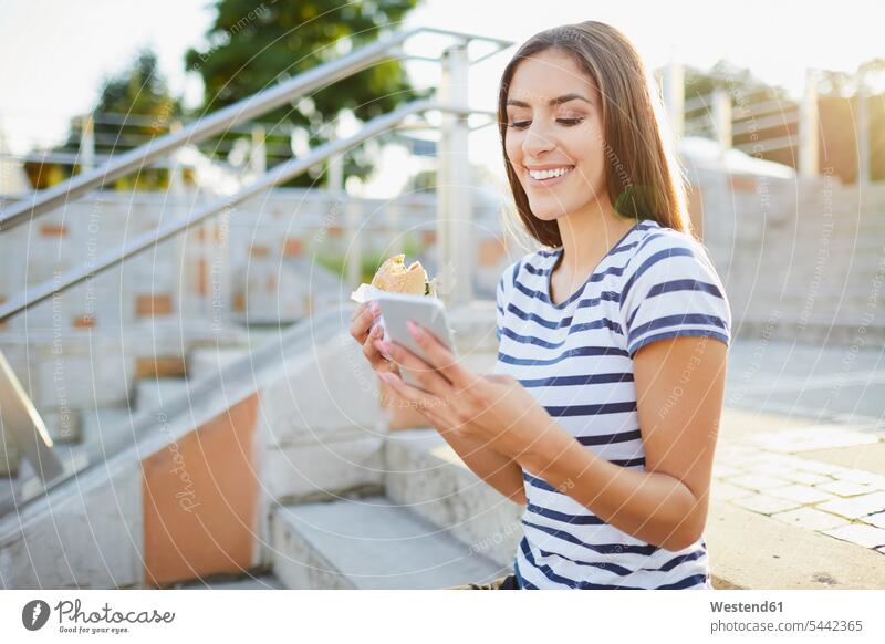 Young woman sitting on stairs eating bagel and using smartphone females women smiling smile mobile phone mobiles mobile phones Cellphone cell phone cell phones