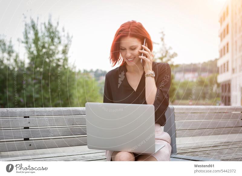 Young woman sitting on bench with her laptop, making a phone call females women smart smart casual smart-casual Business Casual well dressed young Smartphone