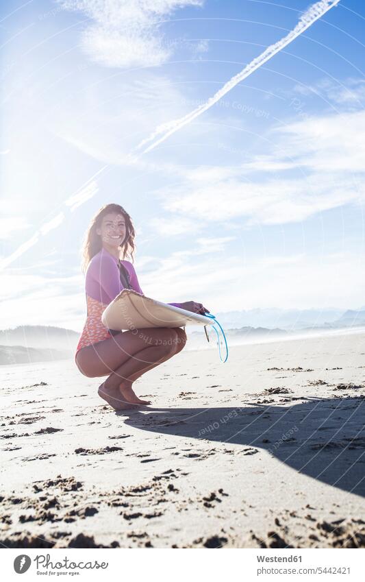 Smiling woman with surfboard crouching on the beach beaches smiling smile surfer female surfer surfers female surfers cowering surfboards females women surfing