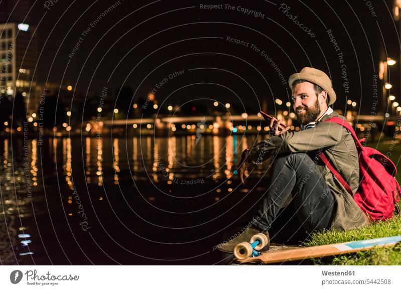 Smiling young man with cell phone and skateboard sitting at urban riverside at night smiling smile by night nite night photography men males Adults grown-ups