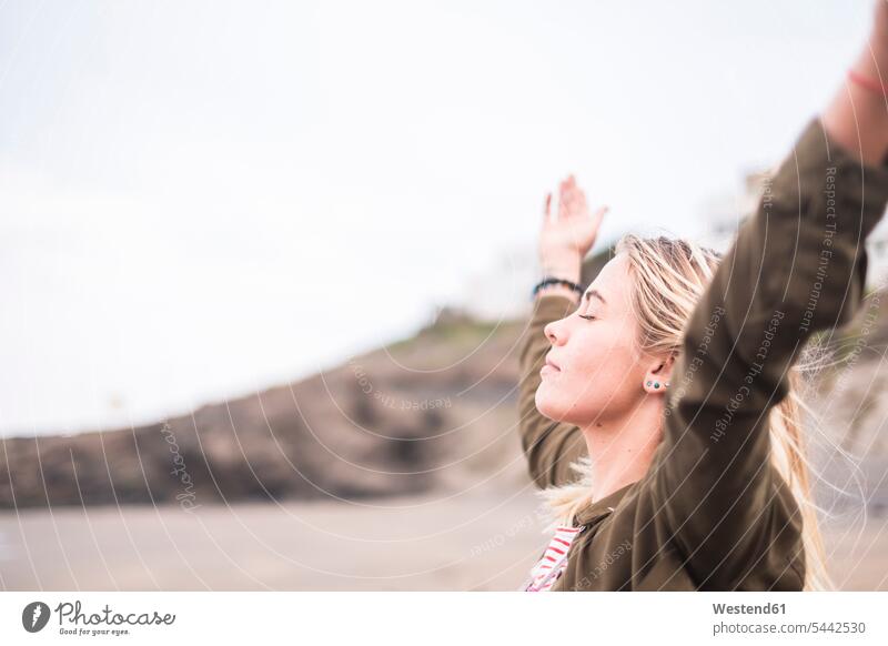 Blond young woman standing on the beach with eyes closed and arms outstretched beaches females women Adults grown-ups grownups adult people persons human being