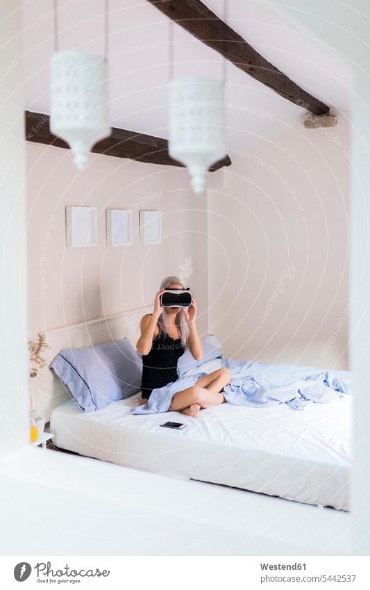 Young woman sitting in bed wearing VR glasses Seated virtual reality specs Eye Glasses spectacles Eyeglasses females women beds Adults grown-ups grownups adult