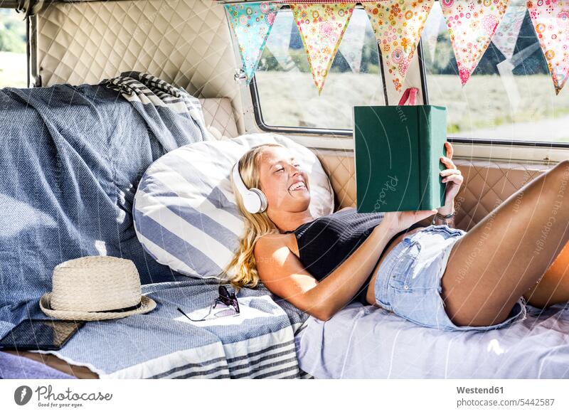 Happy woman with headphones lying in a van reading book books laughing Laughter females women positive Emotion Feeling Feelings Sentiments Emotions emotional