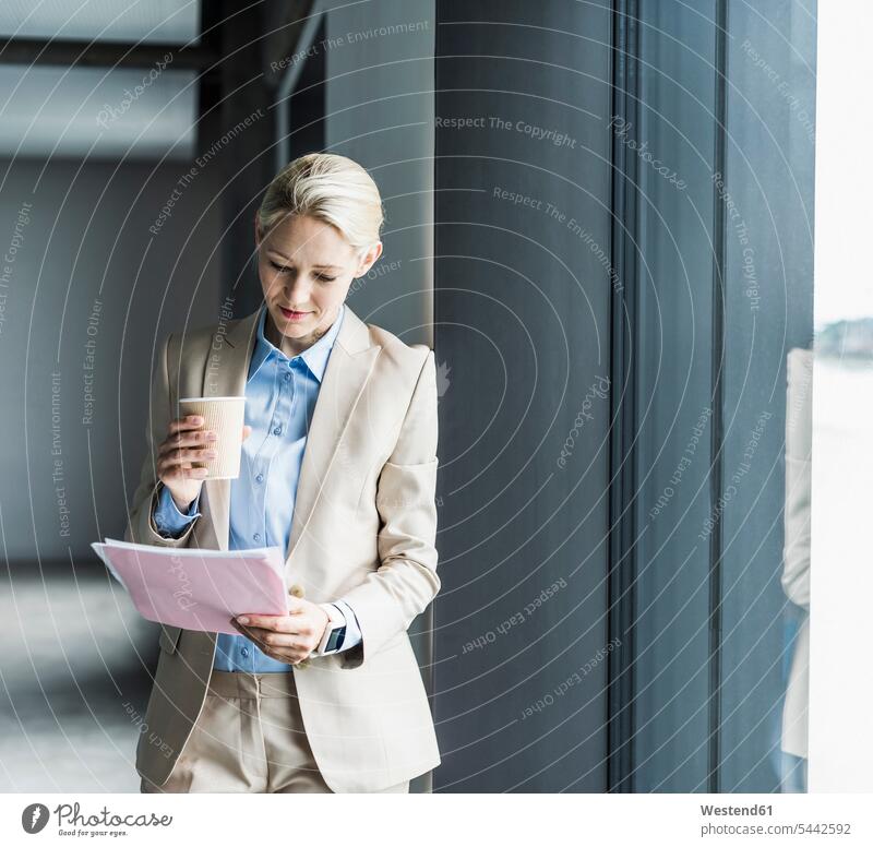 Businesswoman with coffee reading document at the window businesswoman businesswomen business woman business women standing business people businesspeople