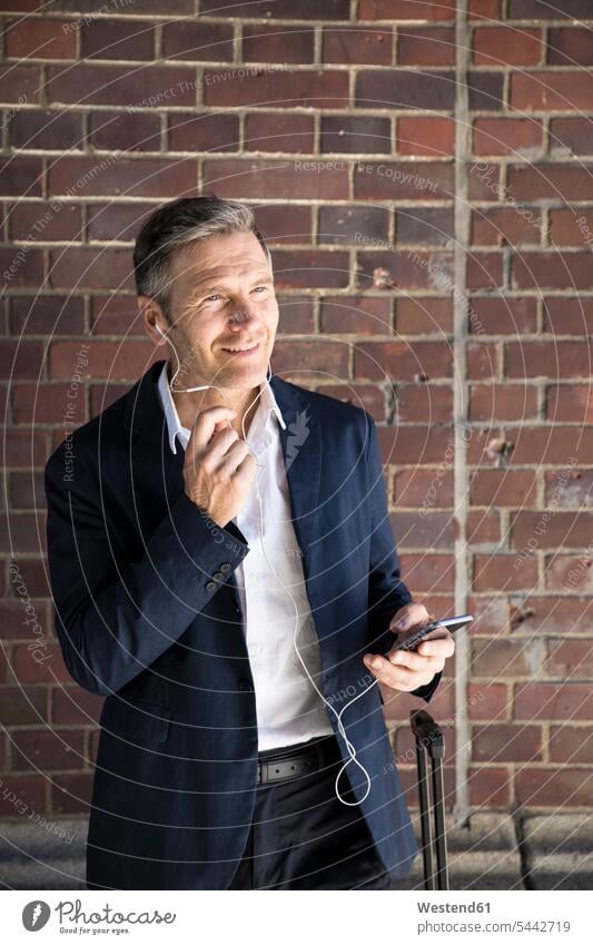 Mature businessman with earphones and smartphone at brick wall ear phone ear phones mobile phone mobiles mobile phones Cellphone cell phone cell phones smiling