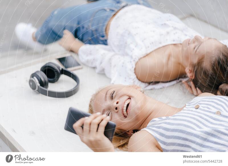 Two happy young women with cell phone and headphones in a skatepark female friends mobile phone mobiles mobile phones Cellphone cell phones Skateboard Park