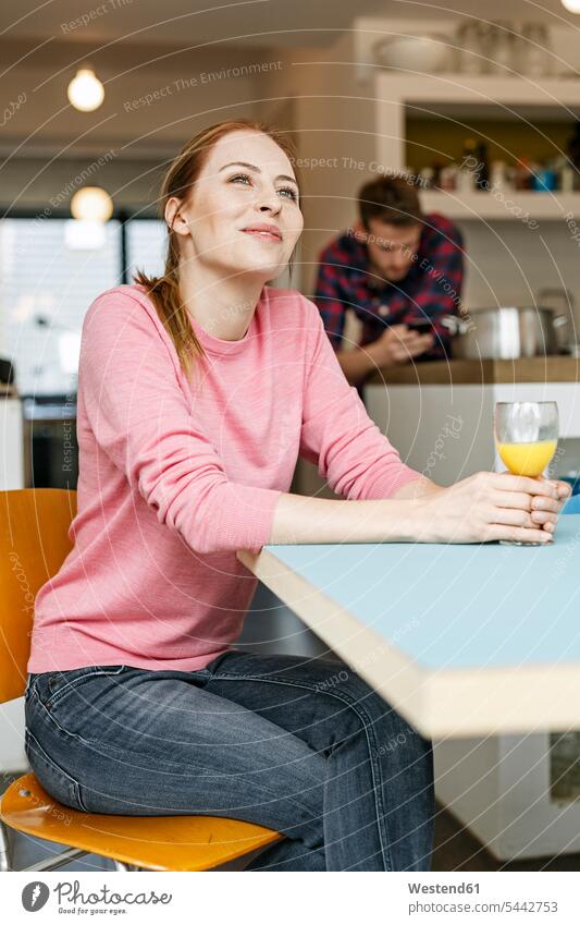 Smiling young woman with glass of orange juice in kitchen at home with man in background females women domestic kitchen kitchens smiling smile Glass