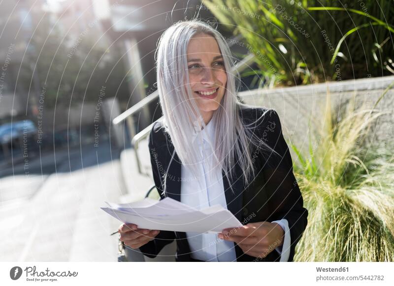 Smiling young businesswoman sitting on a wall in the city with documents Seated paper papers females women smiling smile businesswomen business woman