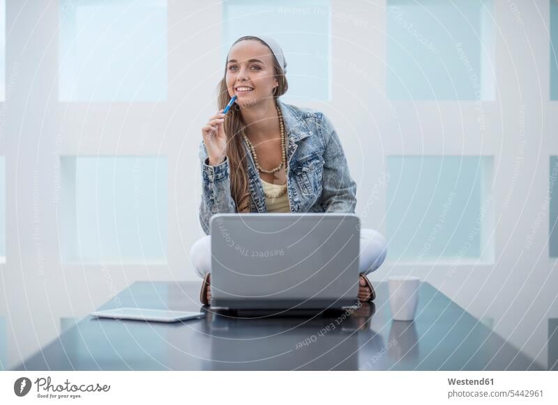 Young woman working in office sitting cross-legged on desk with her laptop office worker happiness happy At Work Joy enjoyment pleasure Pleasant delight