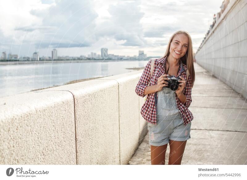 Smiling young woman with a camera at the riverside cameras smiling smile portrait portraits females women River Rivers Adults grown-ups grownups adult people