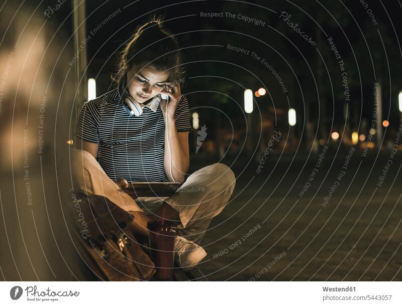 Young woman with smoothie sitting on bench at night using tablet and headphones by night nite night photography females women Adults grown-ups grownups adult