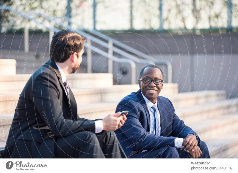 Two businessmen sitting on stairs talking speaking colleagues Businessman Business man Businessmen Business men smiling smile stairway business people