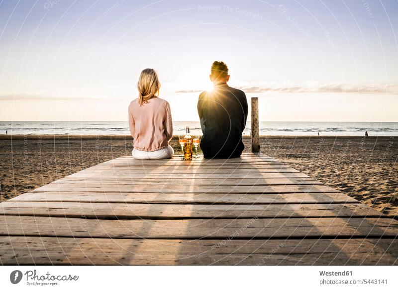 Couple with wine glasses sitting on boardwalk on the beach at sunset beaches relaxed relaxation Seated Wine couple twosomes partnership couples relaxing Alcohol