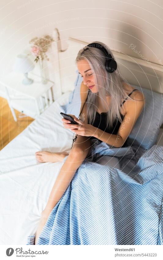 Young woman in bed with cell phone and headphones beds headset females women mobile phone mobiles mobile phones Cellphone cell phones Adults grown-ups grownups
