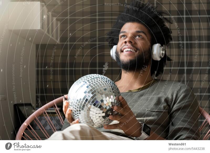 Man sitting in living room in armchair holding mirror ball listening to music man men males smiling smile hearing Seated headphones headset relaxed relaxation
