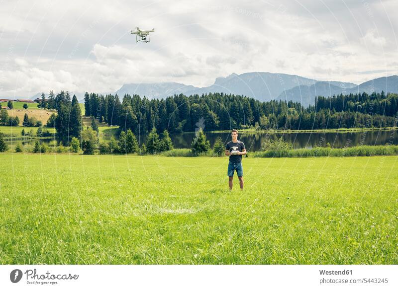 Germany, Bavaria, Pfronten, young man flying his drone at lakeside View Vista Look-Out outlook drones men males standing meadow meadows Adults grown-ups