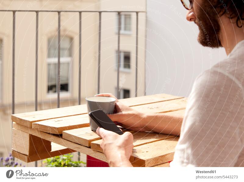 Man sitting with coffee mug on balcony using cell phone balconies man men males Smartphone iPhone Smartphones Adults grown-ups grownups adult people persons